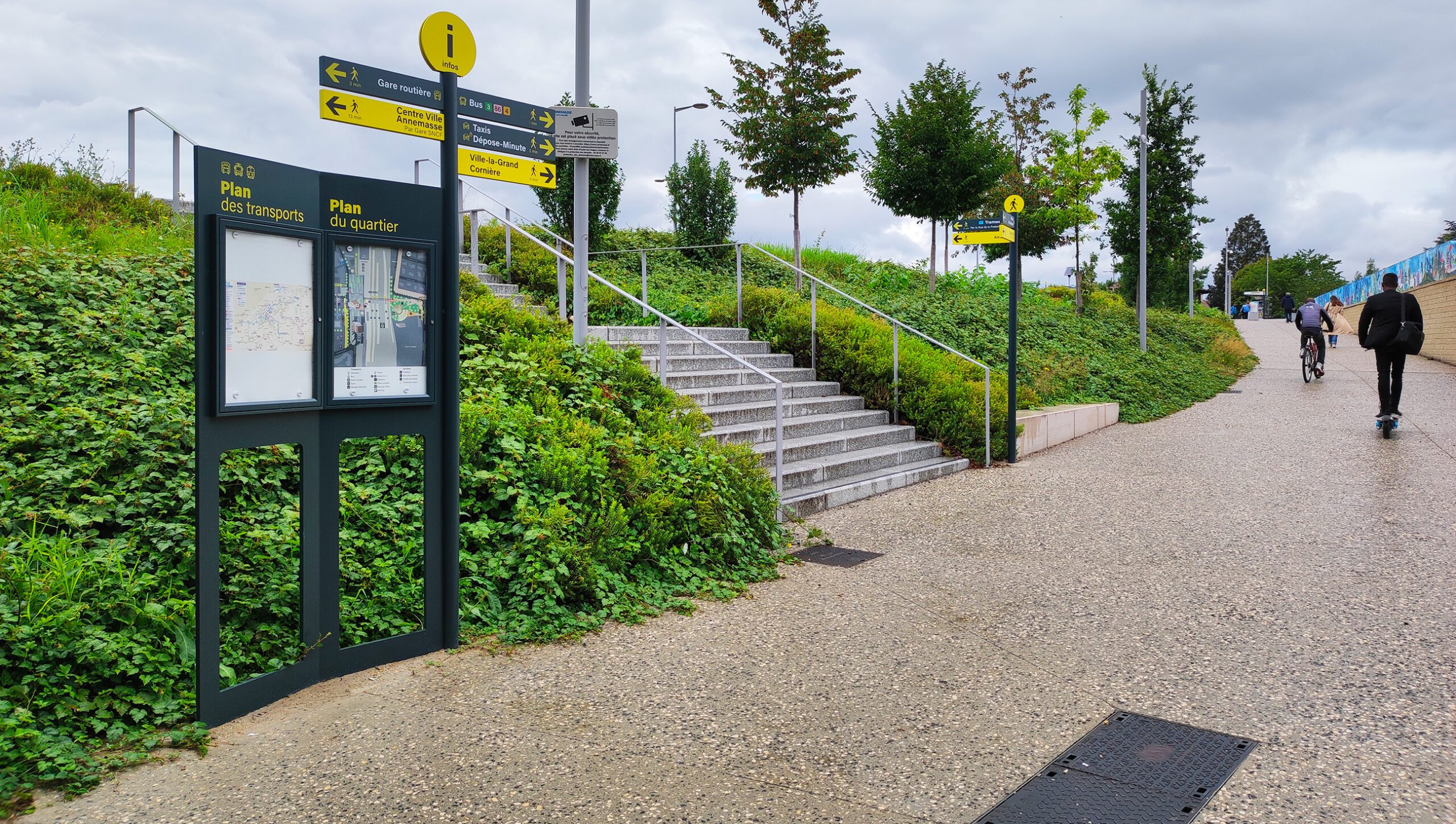Totem avec plan des transports et du quartier, escaliers et panneaux directionnels sur un chemin piéton arboré
