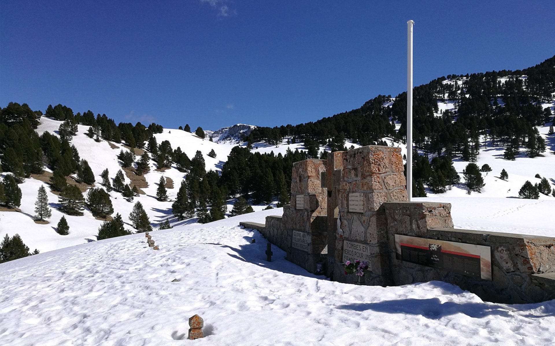 Monument commémoratif en pierre dans une nécropole enneigée de montagne, avec sapins et drapeau blanc
