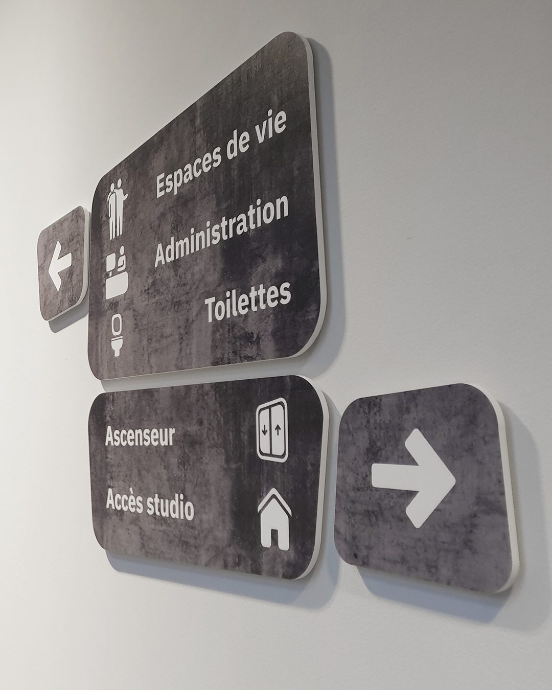 Panneaux directionnels sur mur blanc indiquant Espaces de vie, Administration, Toilettes, Ascenseur et Accès studio