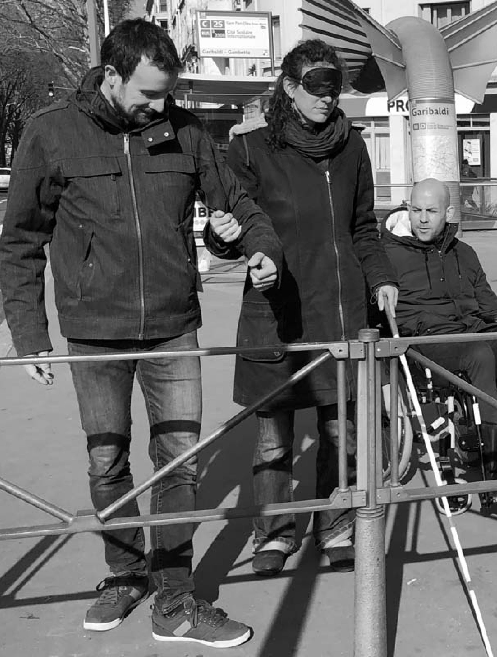 Deux personnes debout et une en fauteuil roulant devant une barrière urbaine près de l'arrêt Garibaldi