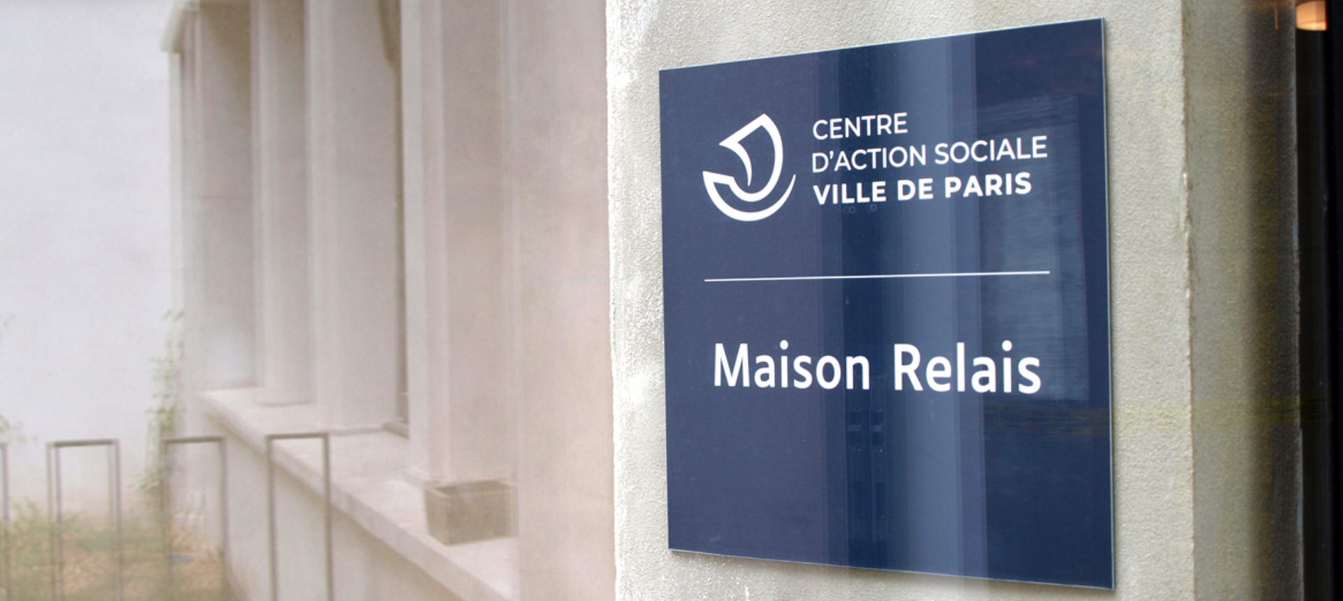 Panneau bleu Centre d’Action Sociale Ville de Paris indiquant Maison Relais fixé sur une façade claire