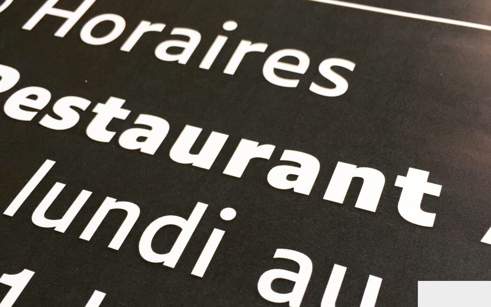 Gros plan en biais sur une signalétique noire à grands caractères blancs, avec les mots « Horaires » et « restaurant » visibles