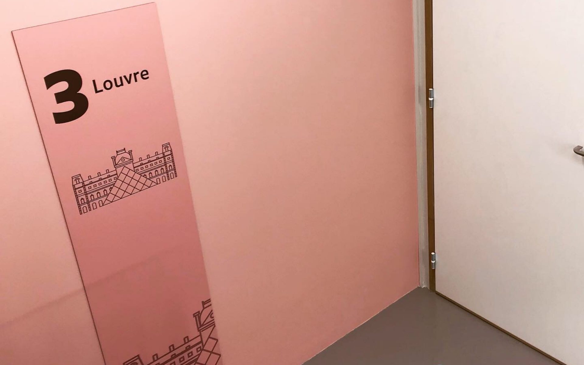 Panneau rose incliné marqué 3 Louvre avec dessin du musée, posé contre un mur près d'une porte blanche