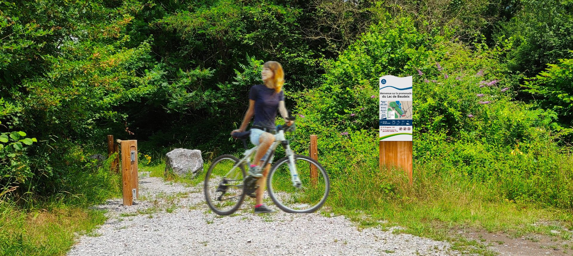 Cycliste sur un chemin gravillonné bordé de végétation, près d'un panneau du lac de Baudreix