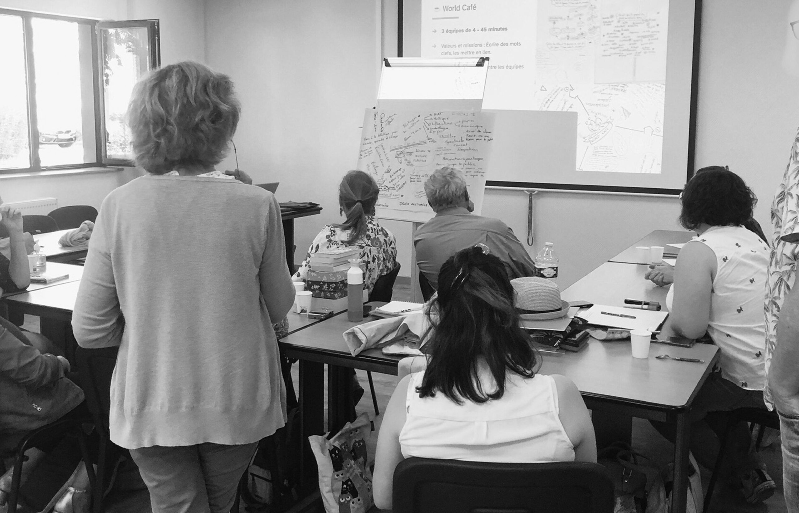 Participants en atelier créatif assis autour de tables face à un paperboard et une projection World Café