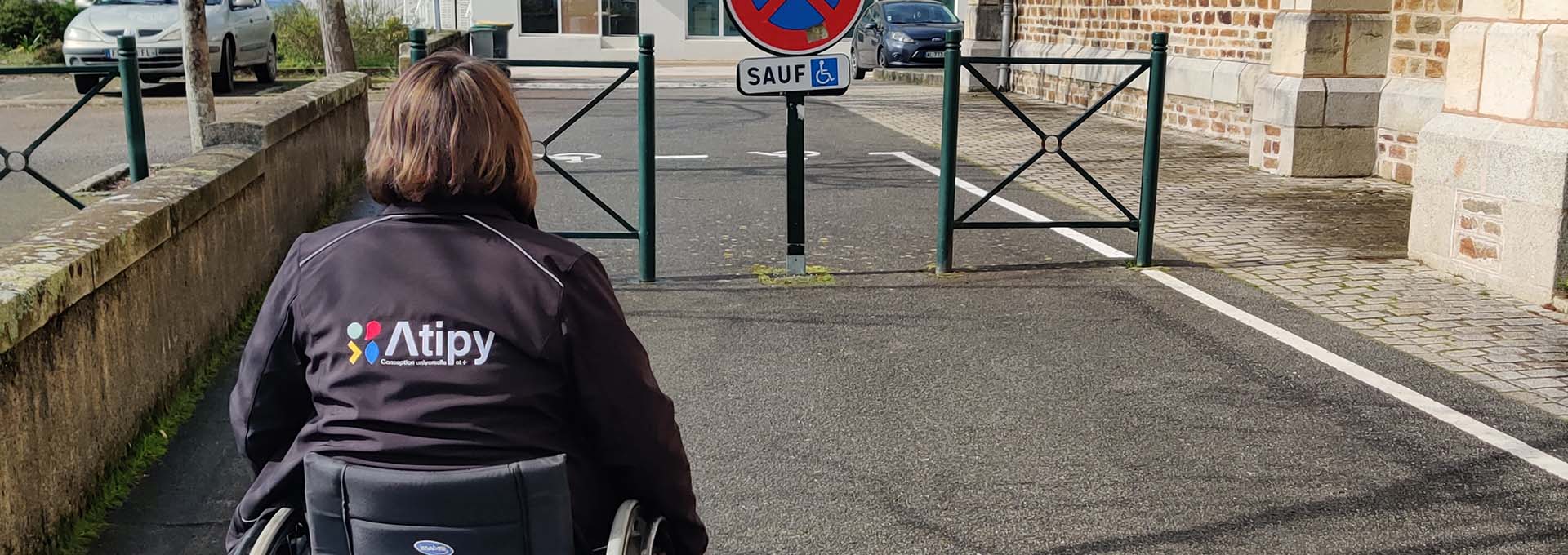 Personne en fauteuil roulant avec veste Atipy face à un accès barré et un panneau « Sauf » avec symbole PMR