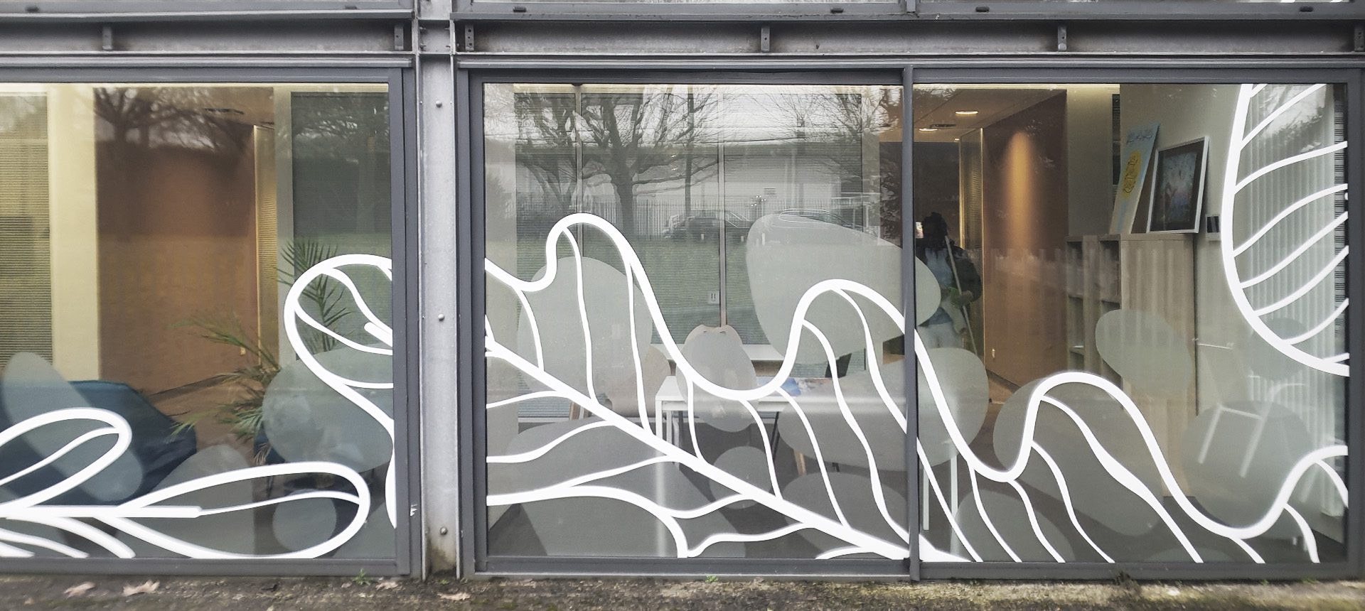 Baies vitrées d’un bureau avec motifs blancs de feuilles, laissant voir tables, chaises et accueil intérieur