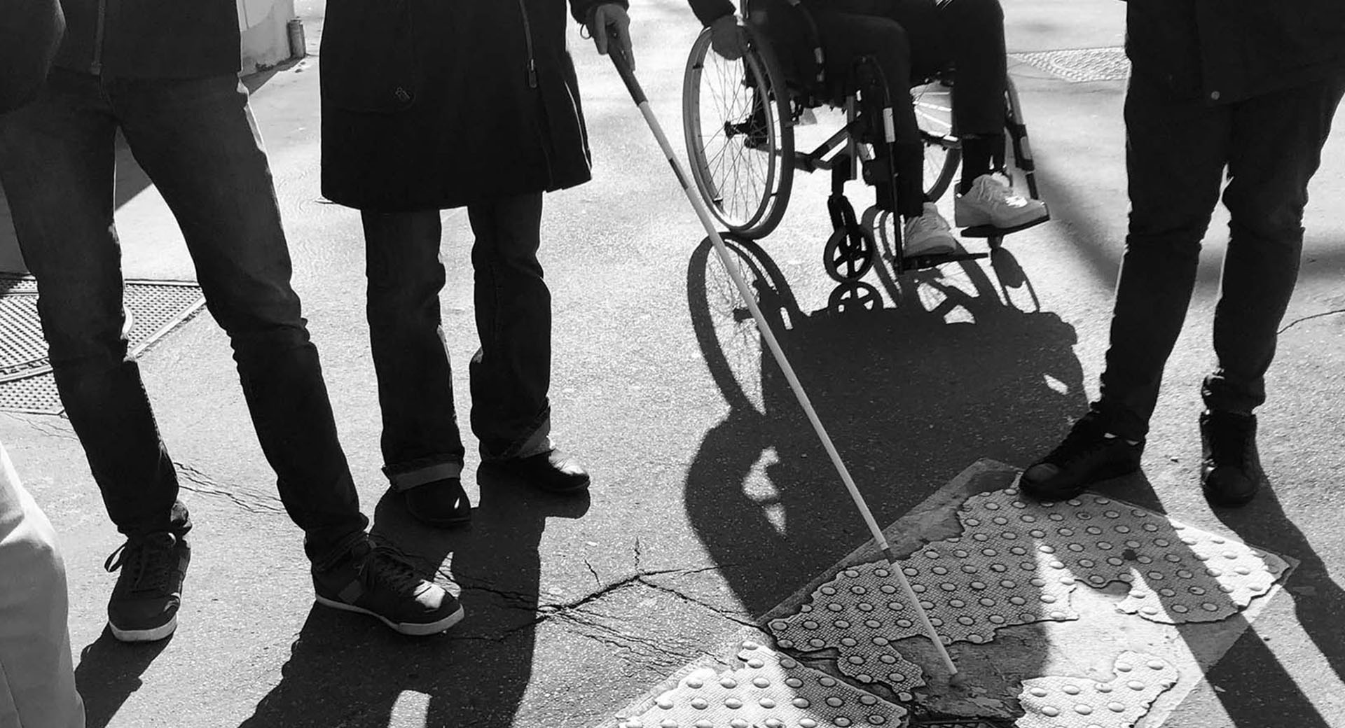Personnes autour d’une dalle podotactile, avec canne blanche et fauteuil roulant sur un trottoir extérieur