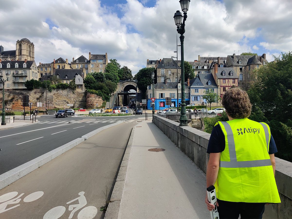 Personne en gilet jaune Atipy marchant sur un pont avec piste cyclable face à un centre-ville ancien