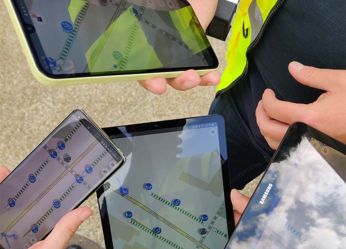 Trois personnes consultent sur tablette et smartphone une carte avec pictogrammes et tracés de relevé urbain