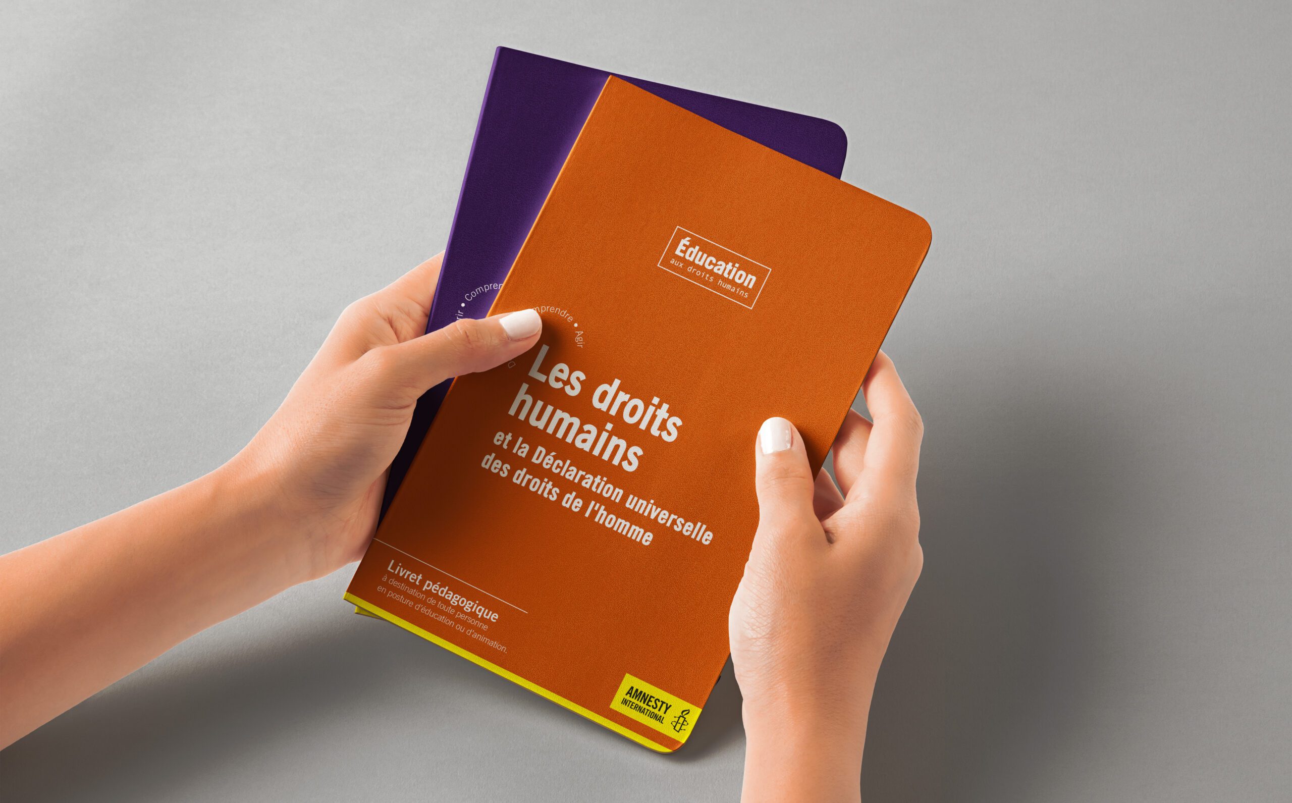 Deux mains tiennent des livrets Amnesty International sur les droits humains, aux couvertures orange et violette