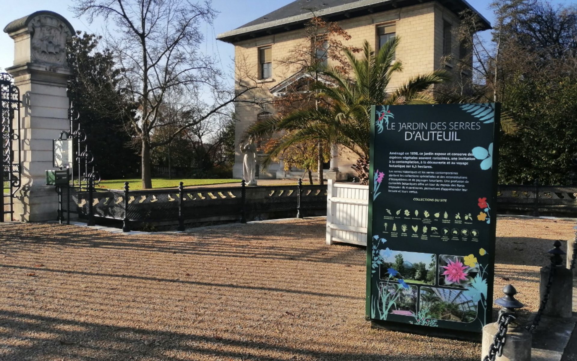 Panneau d’accueil Le Jardin des Serres d’Auteuil devant une bâtisse claire et des palmiers dans le jardin